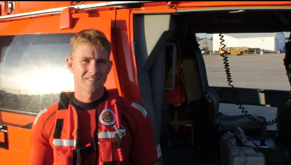 Drew Sinclair stands in front of a Coast Guard helicopter with an orange wetsuit and a red life jacket.