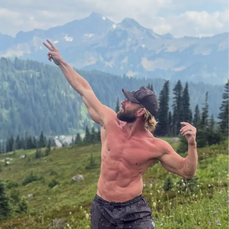 Drew Sinclair stands, flexing, on a green hill in front of mountains, with a lake in a valley below.